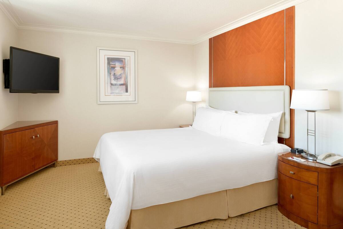 Une chambre d’hôtel avec un lit, une table de chevet en bois, un bureau, un téléviseur à écran plat et un tableau encadré au mur.