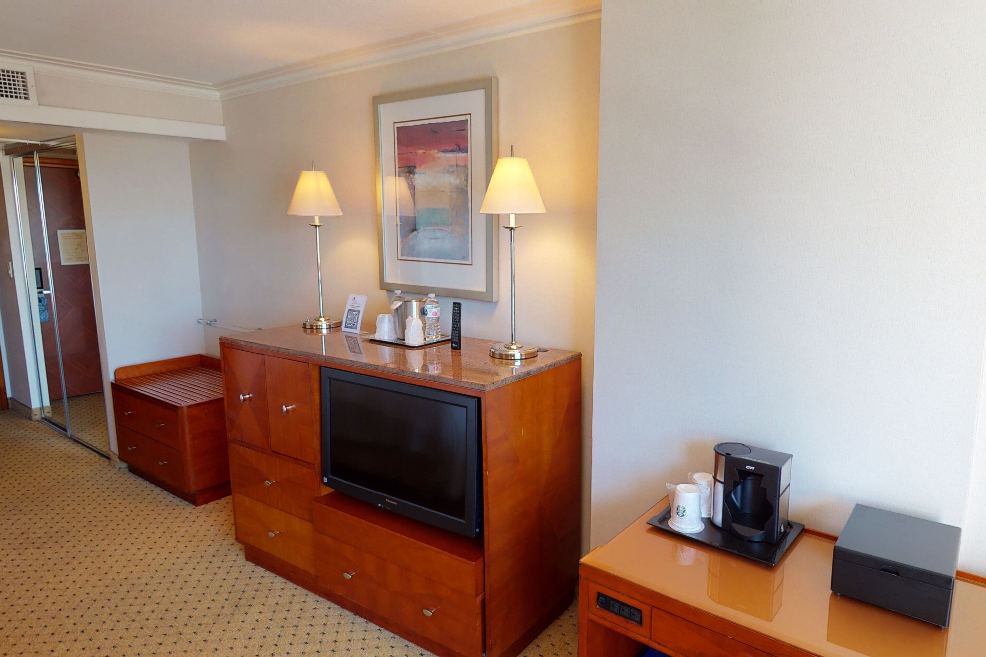 A hotel room with a wooden dresser, a flat-screen TV, a coffee maker, and two lamps.