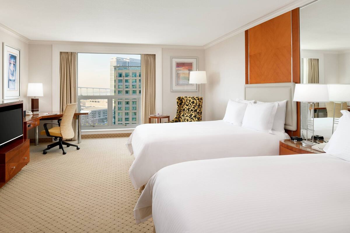 Une chambre d’hôtel avec deux lits, un bureau, une chaise, une télévision et une grande fenêtre donnant sur la ville.