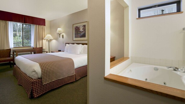 Une chambre d’hôtel dotée d’un grand lit, d’un coin salon avec un fauteuil et une table, d’une salle de bain avec baignoire et d’une fenêtre avec des rideaux.