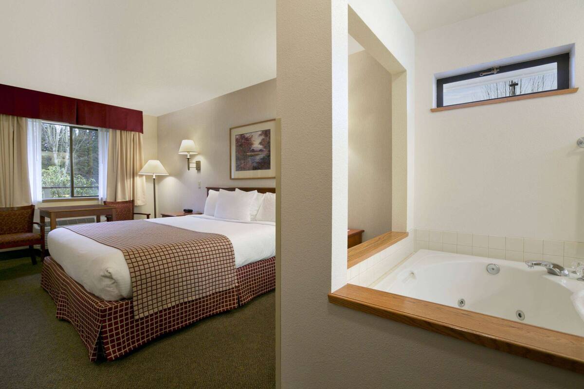 Een hotelkamer met een groot bed, een zithoek met een stoel en tafel, een badkamer met een bad en een raam met gordijnen.