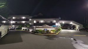L’entrée d’un hôtel de nuit, avec un autobus garé à gauche, plusieurs voitures stationnées sur le parking, et un bâtiment bien éclairé avec une enseigne indiquant le hall d’entrée.