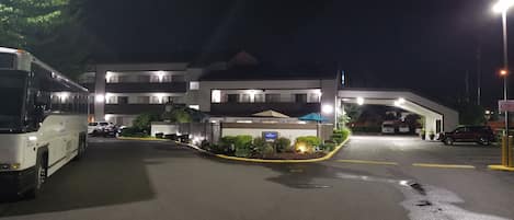 A hotel entrance at night with a bus parked on the left, multiple cars parked in the lot, and a well-lit building with a sign indicating a lobby.