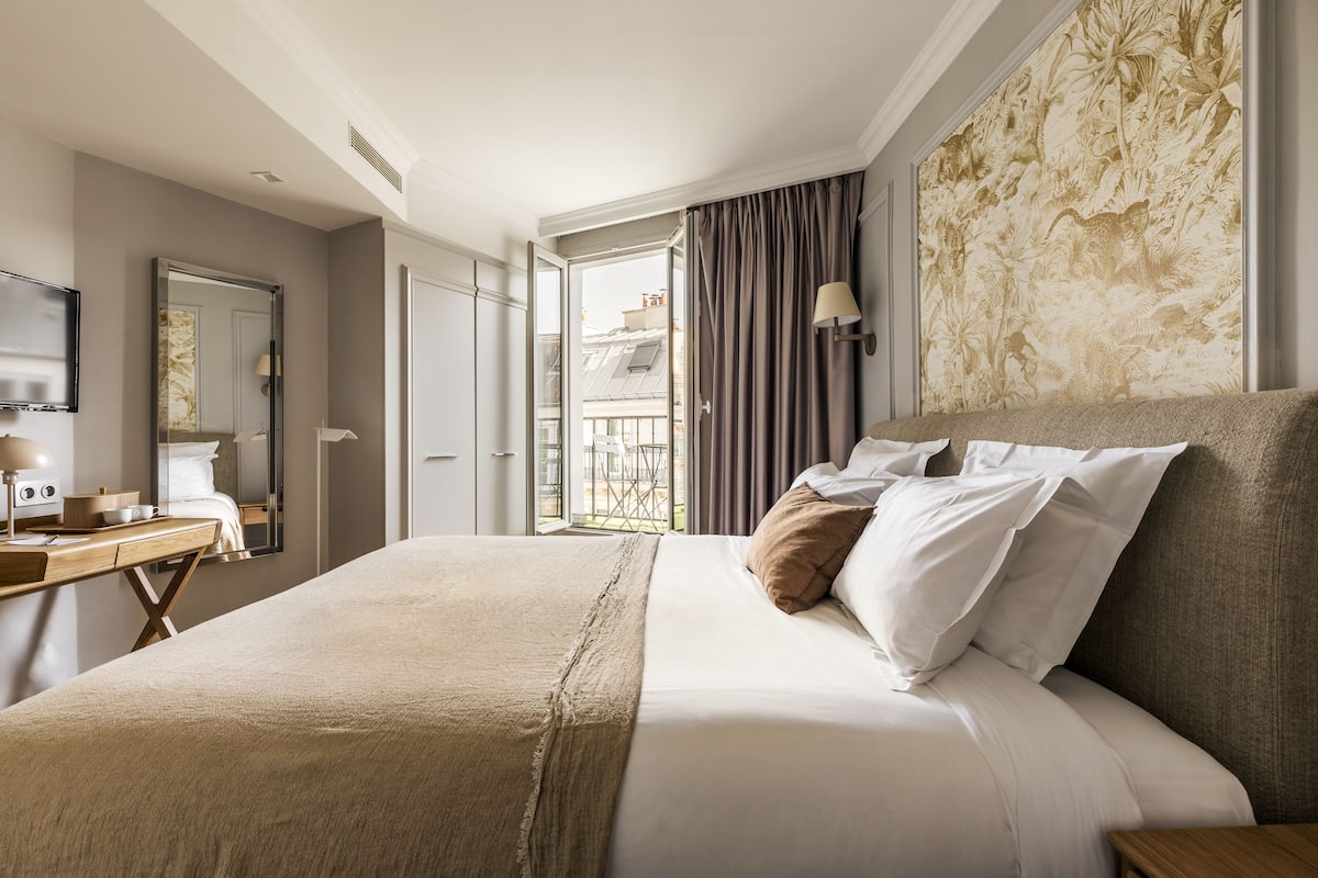 A hotel room with a large bed, a desk with a lamp, and a view of the city through an open door.