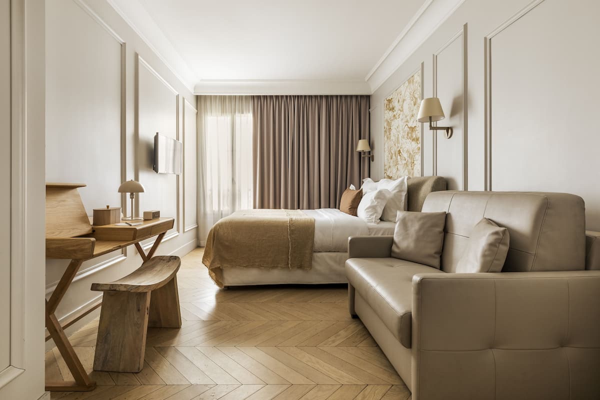 A modern hotel room with a bed, a sofa, a wooden bench, a desk, and a lamp.