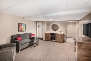 Una habitación de hotel moderna con un sofá gris, una mesita pequeña, un mueble de madera con lavabo y una televisión.