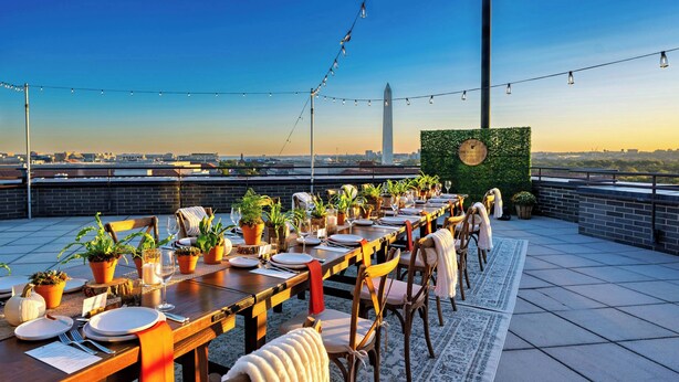 Un área de comedor en la azotea con una larga mesa dispuesta para una comida, plantas en macetas y vistas del horizonte de la ciudad.