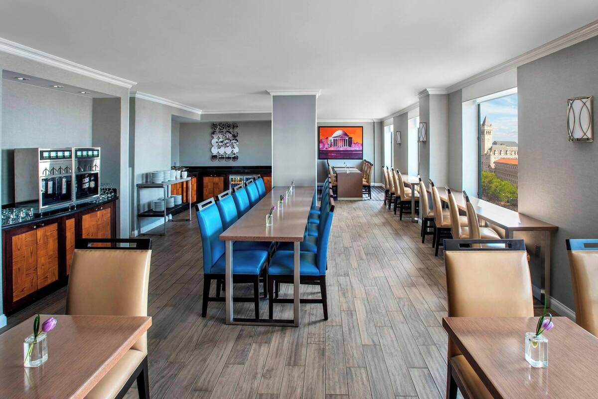 Een modern restaurantgedeelte in een hotel, met een lange gedeelde tafel, blauwe zitbanken en een keukenbar.