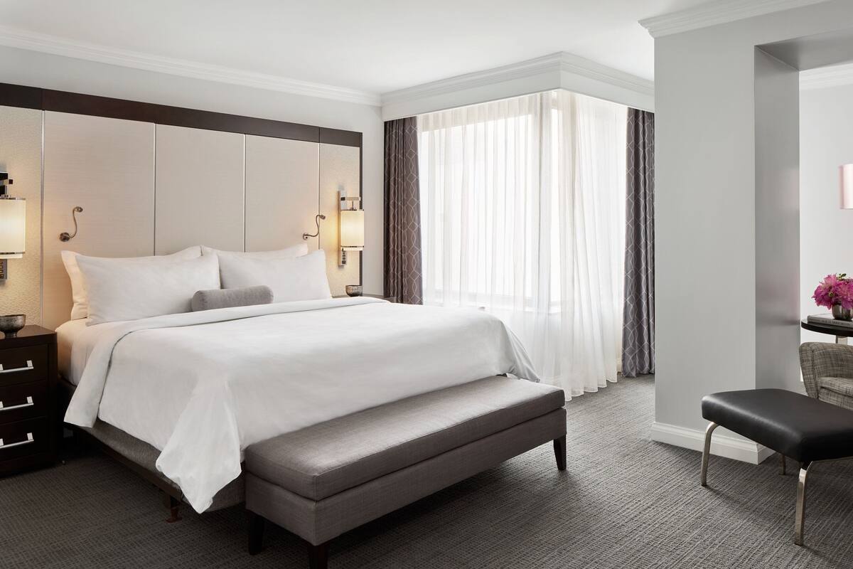 Hotelkamer met een groot bed, een bankje, een bureau met stoel en uitzicht op het buitengebied door dunne gordijnen.