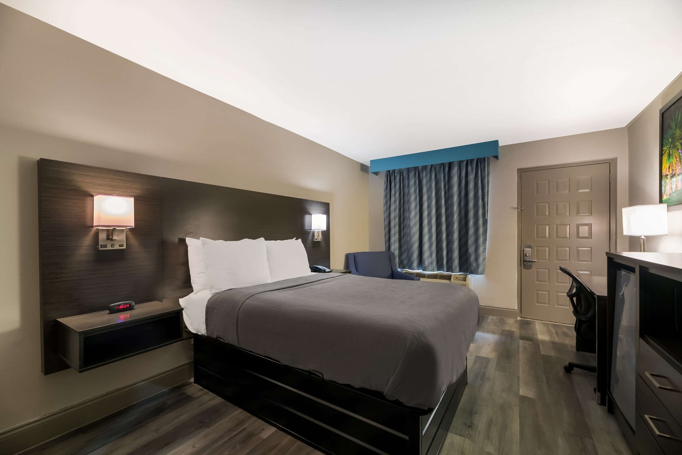 Habitación de hotel con cama, mesitas de noche, escritorio, silla, lámpara de escritorio y puerta.