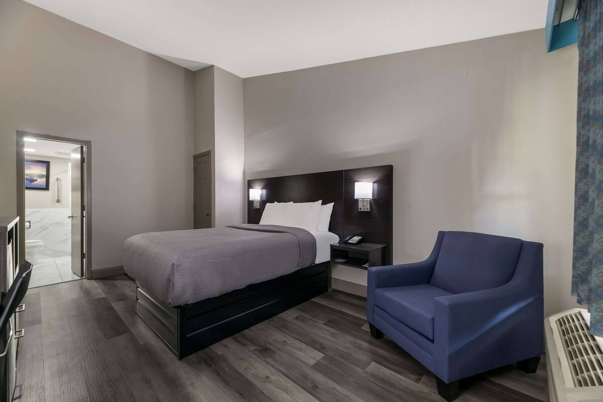Quarto de hotel moderno com uma cama grande, uma poltrona azul, uma mesa de cabeceira com um telefone e um aplique na parede.