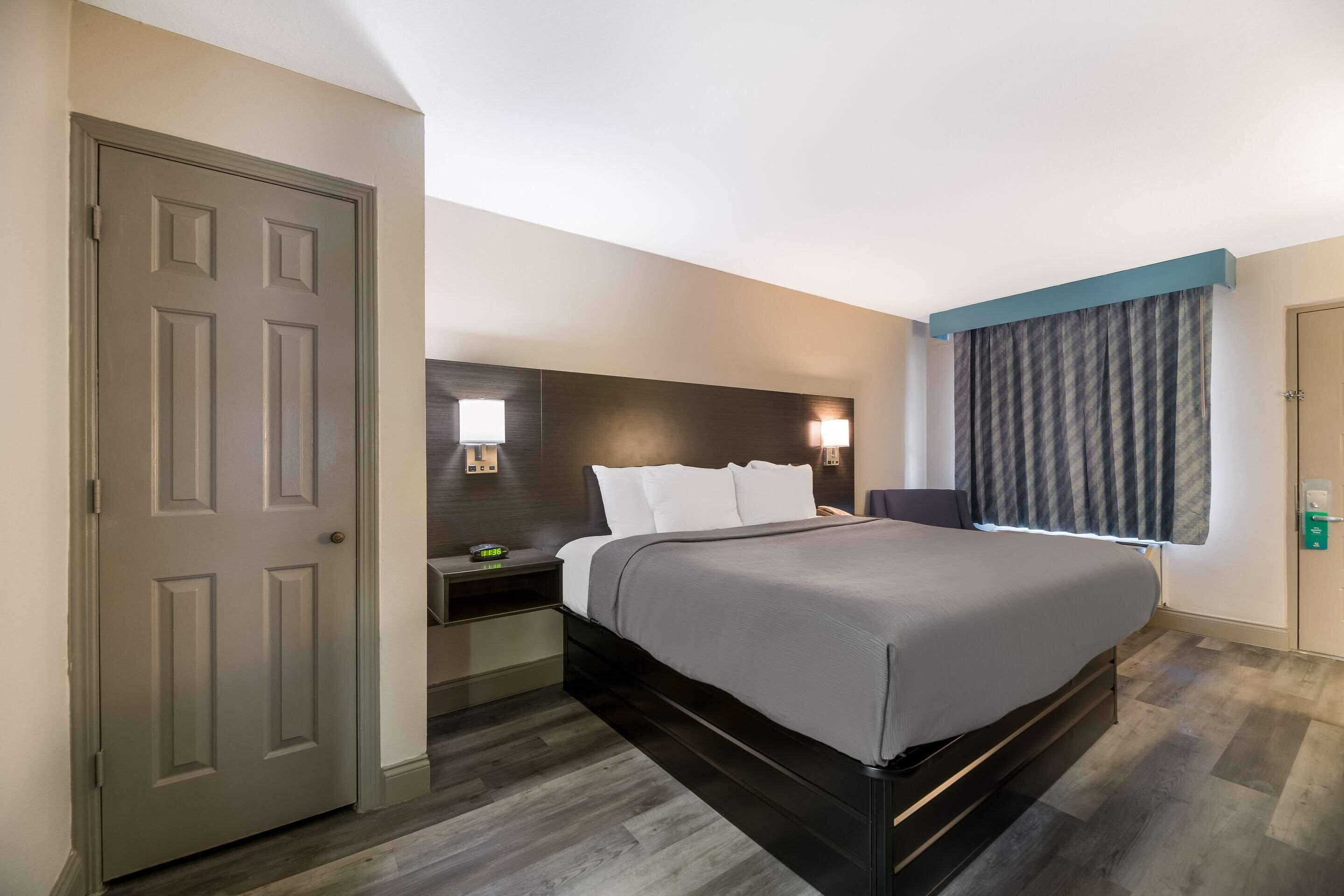 A hotel room with a bed, a nightstand, a lamp, a window with curtains, and a door.