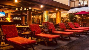 An outdoor seating area with wooden chairs and tables, a bar in the background, and a television screen. - Castle Bamboo Waikīkī Hotel (Honolulu)
