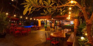 An outdoor dining area with wooden tables and chairs, surrounded by tropical plants and string lights. - Castle Bamboo Waikīkī Hotel (Honolulu)