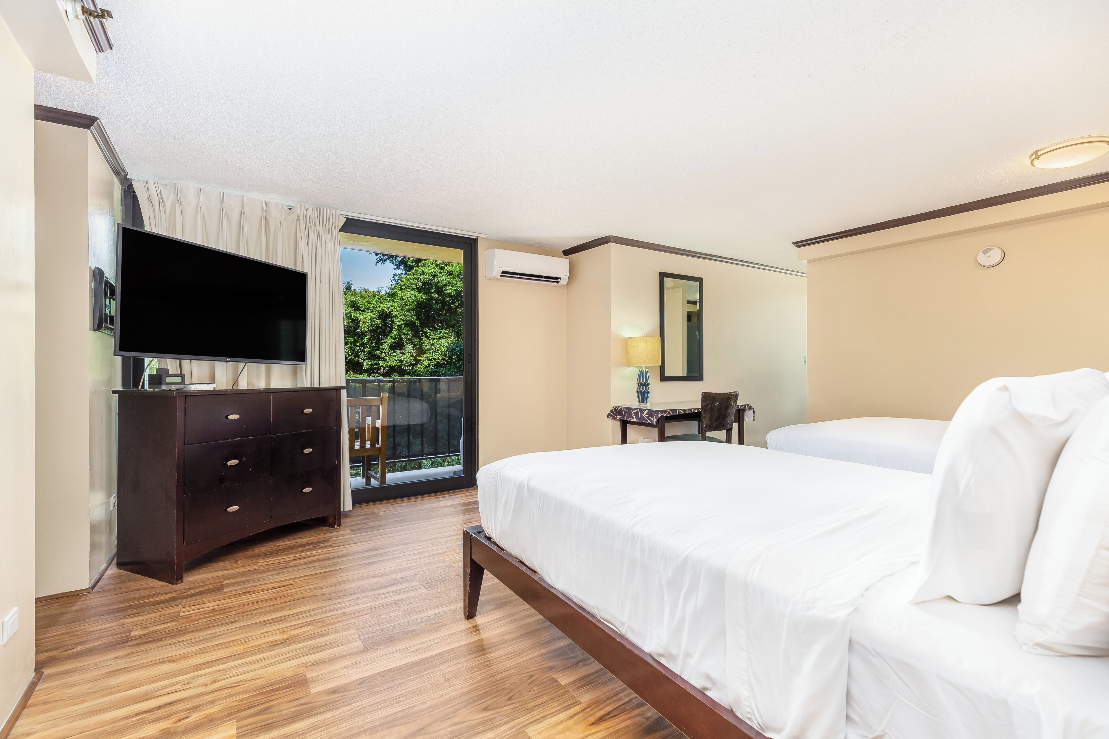A hotel room with a large bed, a flat-screen TV mounted on the wall, a wooden dresser, and a balcony with a view of greenery.