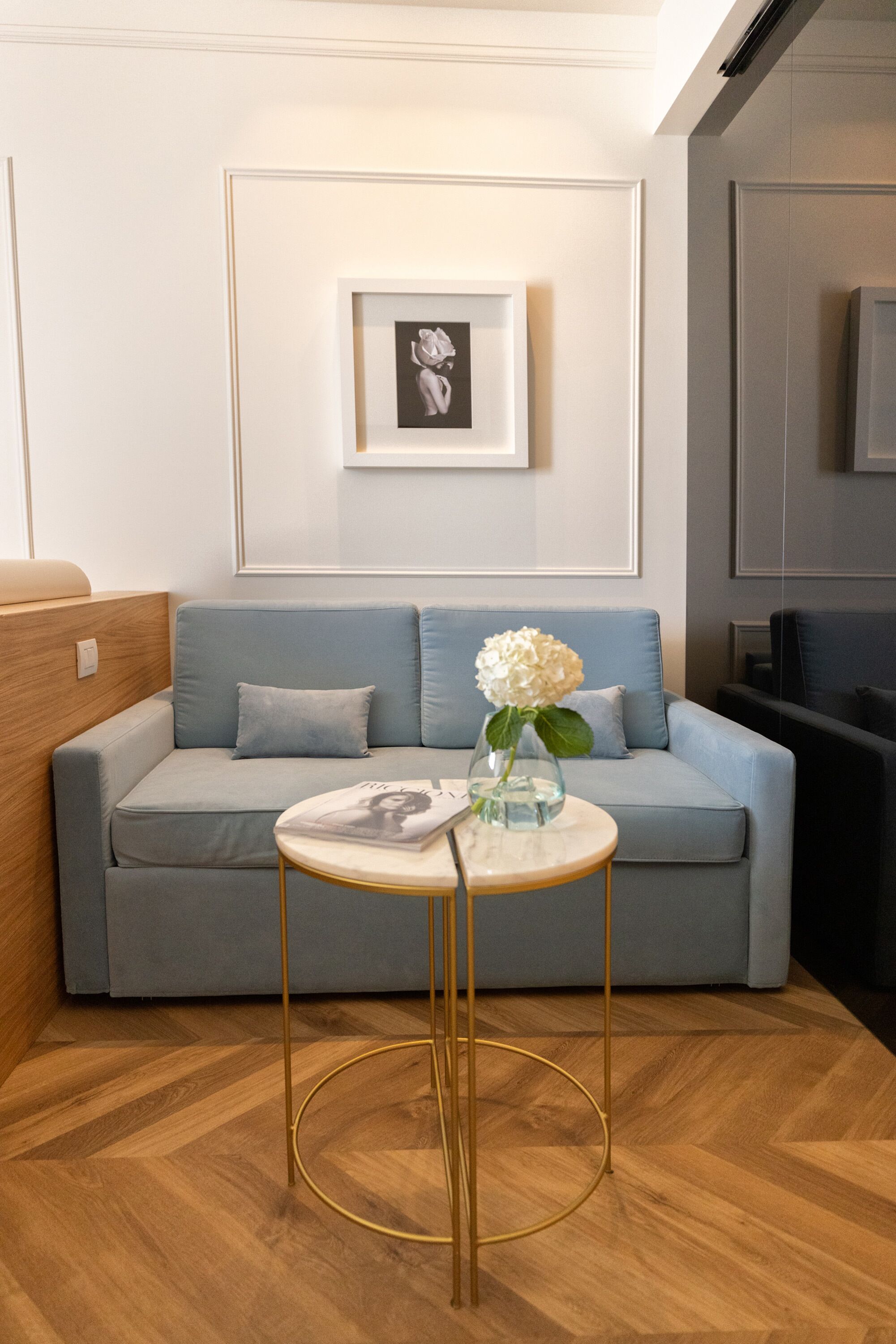 A modern living room with a light blue sofa, a round table with a vase of flowers, and a framed picture on the wall.
