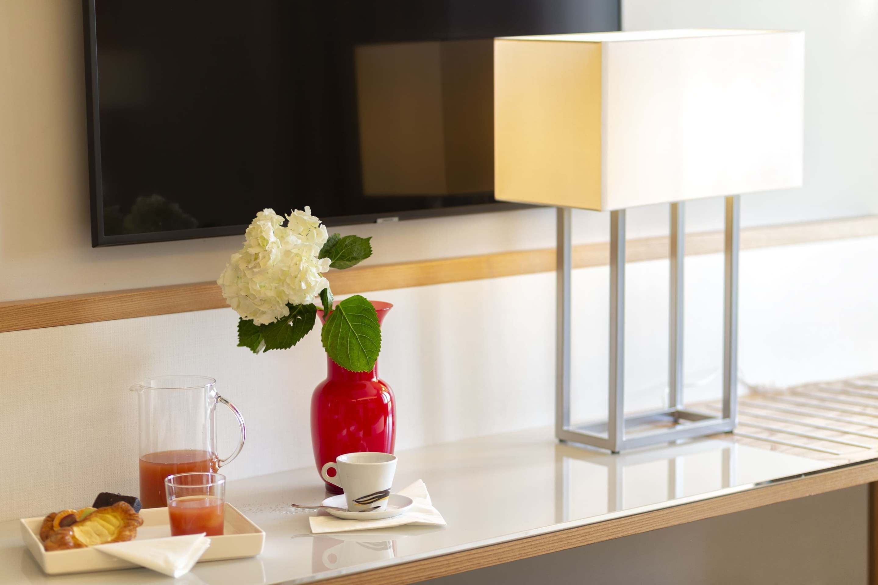 Habitación de hotel con televisión de pantalla plana en la pared, mesita de noche con un jarrón rojo, una taza blanca, una taza de té y una bandeja con pasteles.