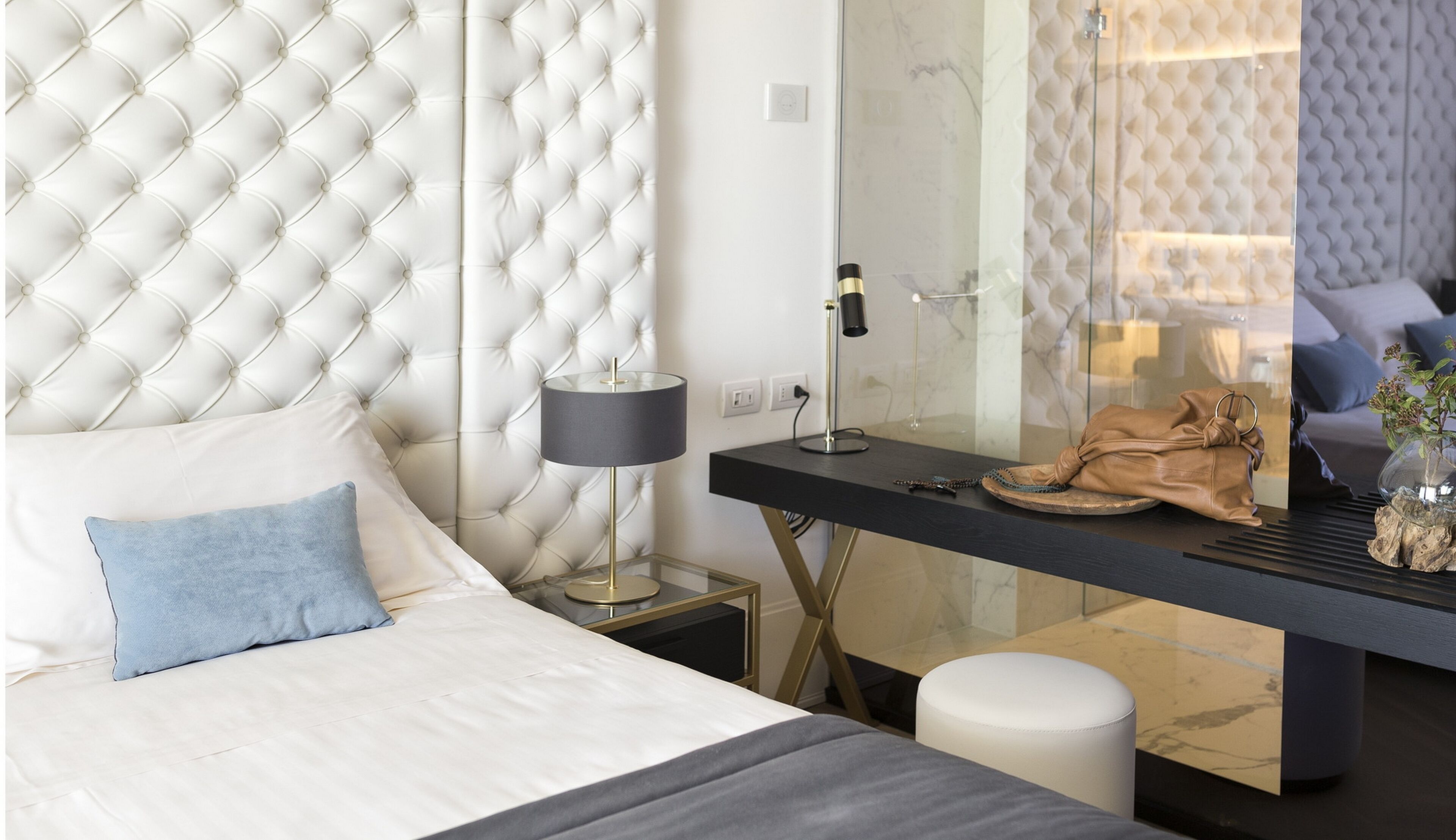 A modern bedroom with a tufted headboard, a bedside table with a lamp, a glass partition, and a view of a living area.