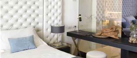 A modern bedroom with a tufted headboard, a bedside table with a lamp, a glass partition, and a view of a living area.