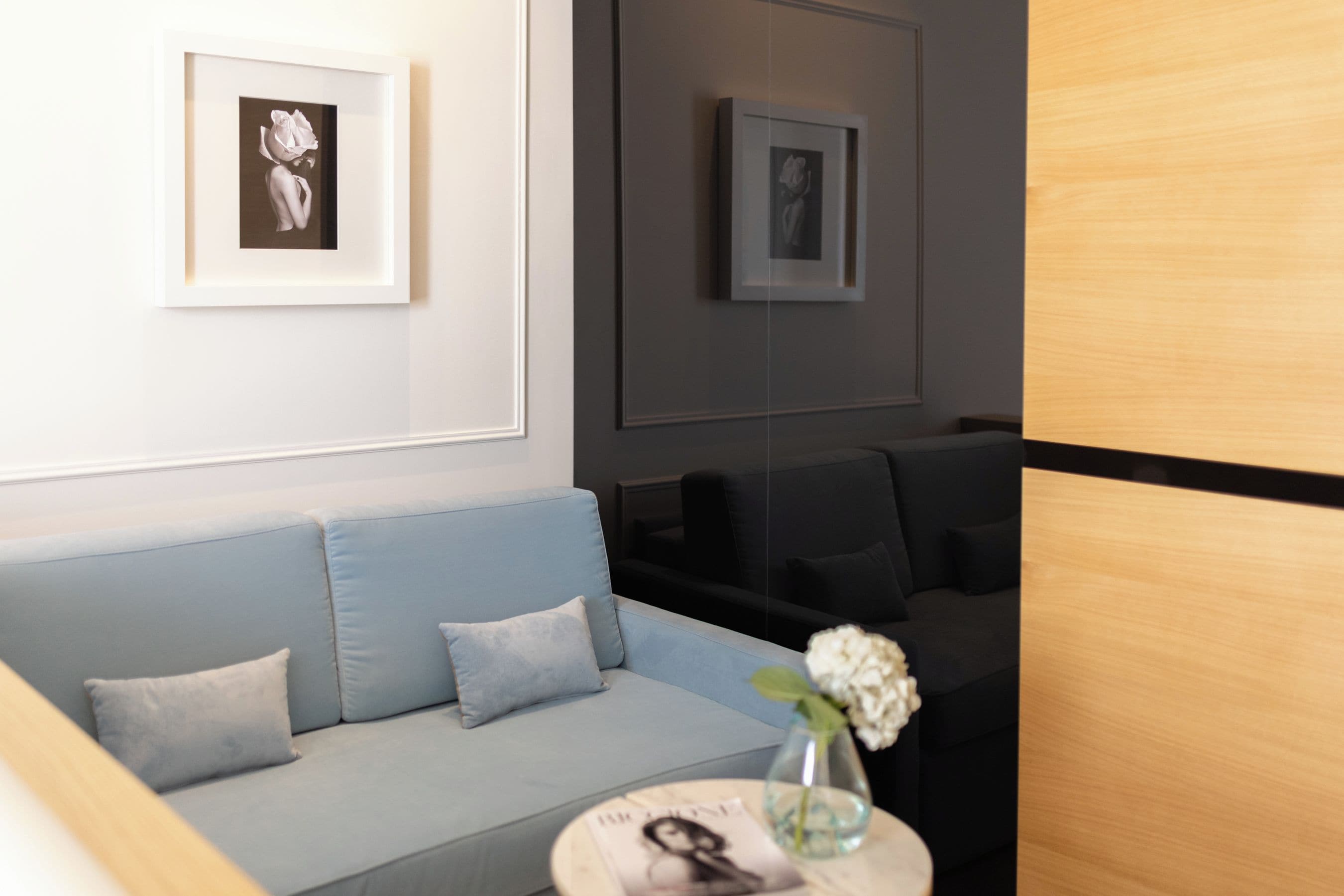 A modern waiting area with a light blue sofa, a small round table with a vase of flowers, and framed artwork on the walls.