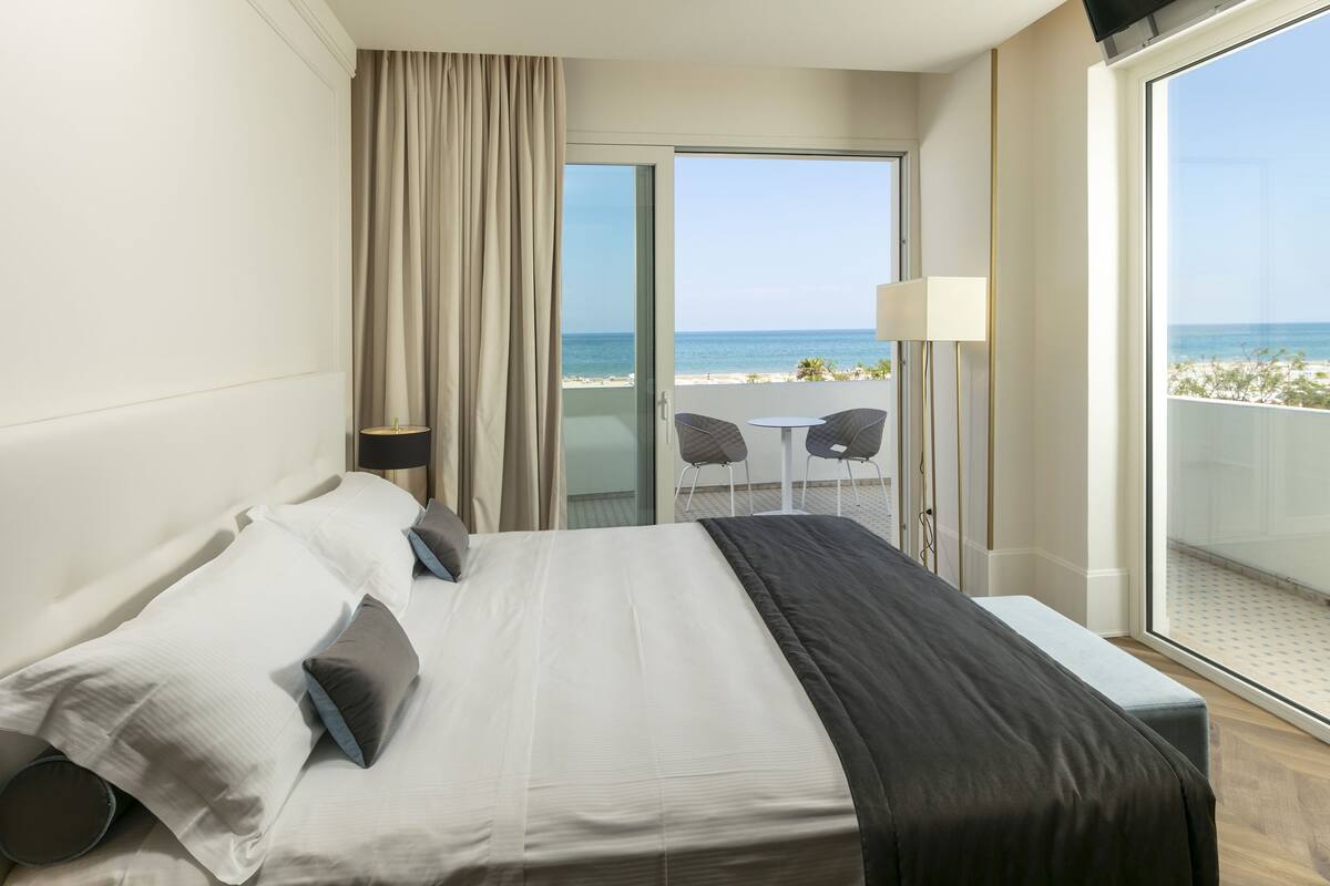 A hotel room with a bed, a window with a view of the beach, a balcony with a table and chairs, and a sliding door leading to the beach.