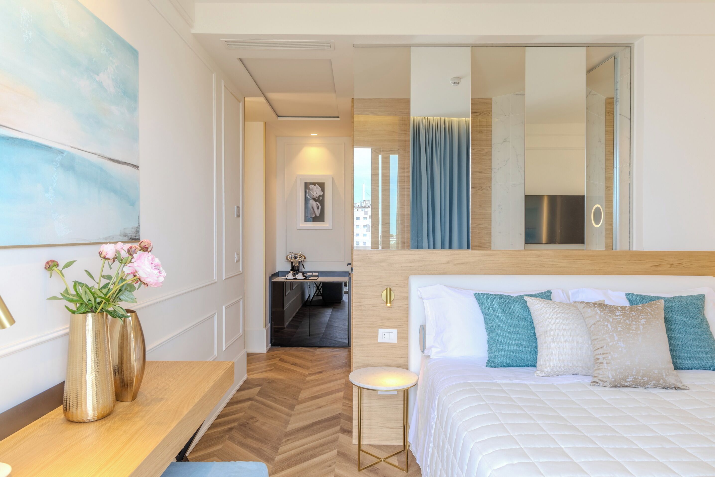 A hotel room with a bed, a desk, a chair, a vase with flowers, a mirror, and a view of the city.