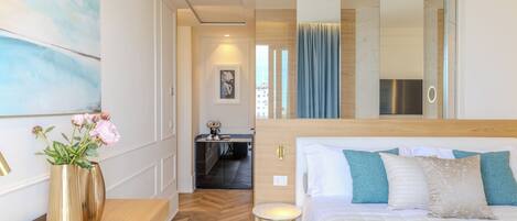 A hotel room with a bed, a desk, a chair, a vase with flowers, a mirror, and a view of the city.