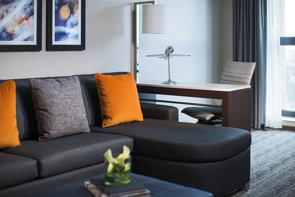 A modern hotel room with a dark grey sectional sofa, a small side table, a desk with a lamp, and a chair.