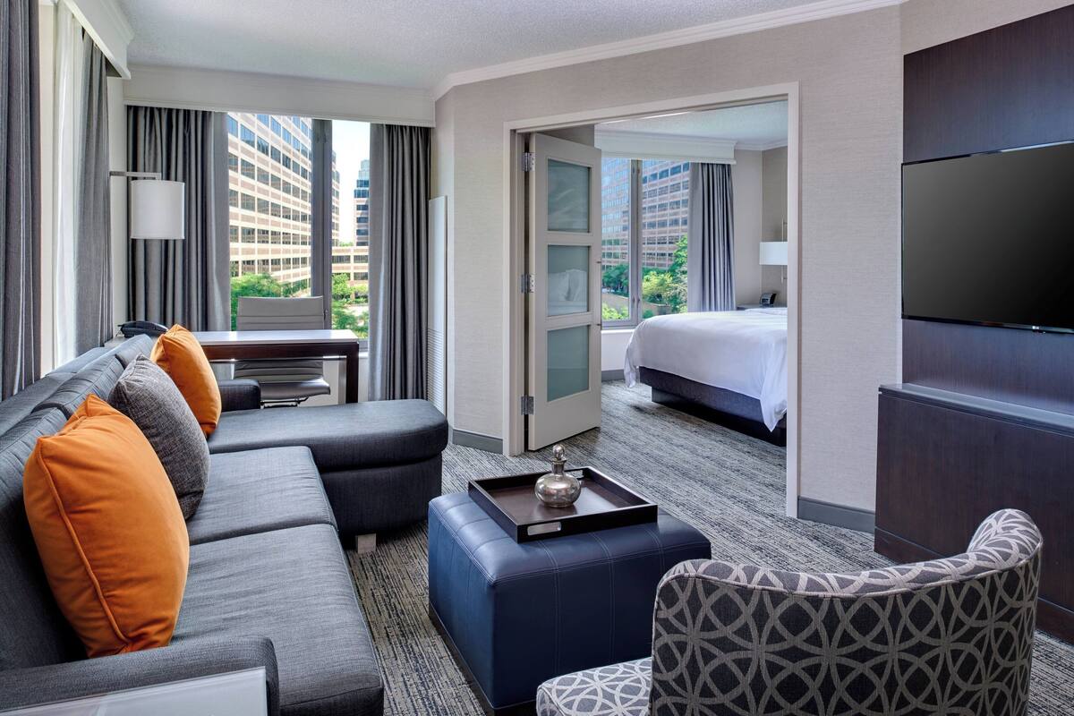 A hotel room with a sofa, ottoman, patterned armchair, a flat-screen TV, a desk with a lamp, and a bed with a view of the cityscape.
