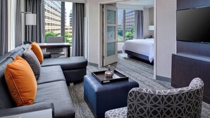 A hotel room with a sofa, ottoman, patterned armchair, a flat-screen TV, a desk with a lamp, and a bed with a view of the cityscape.