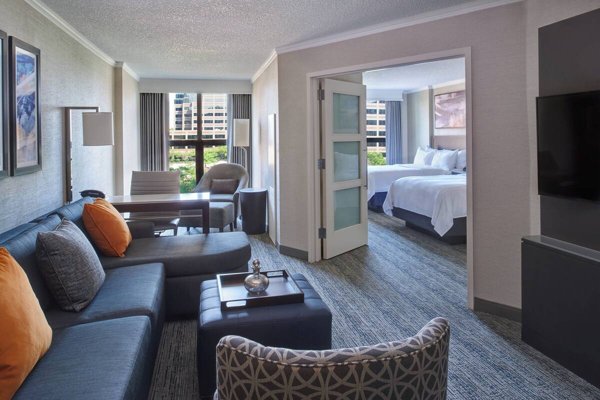 A hotel room with a sofa, armchair, and a bed. There is a TV on the wall and a window with a view of buildings.