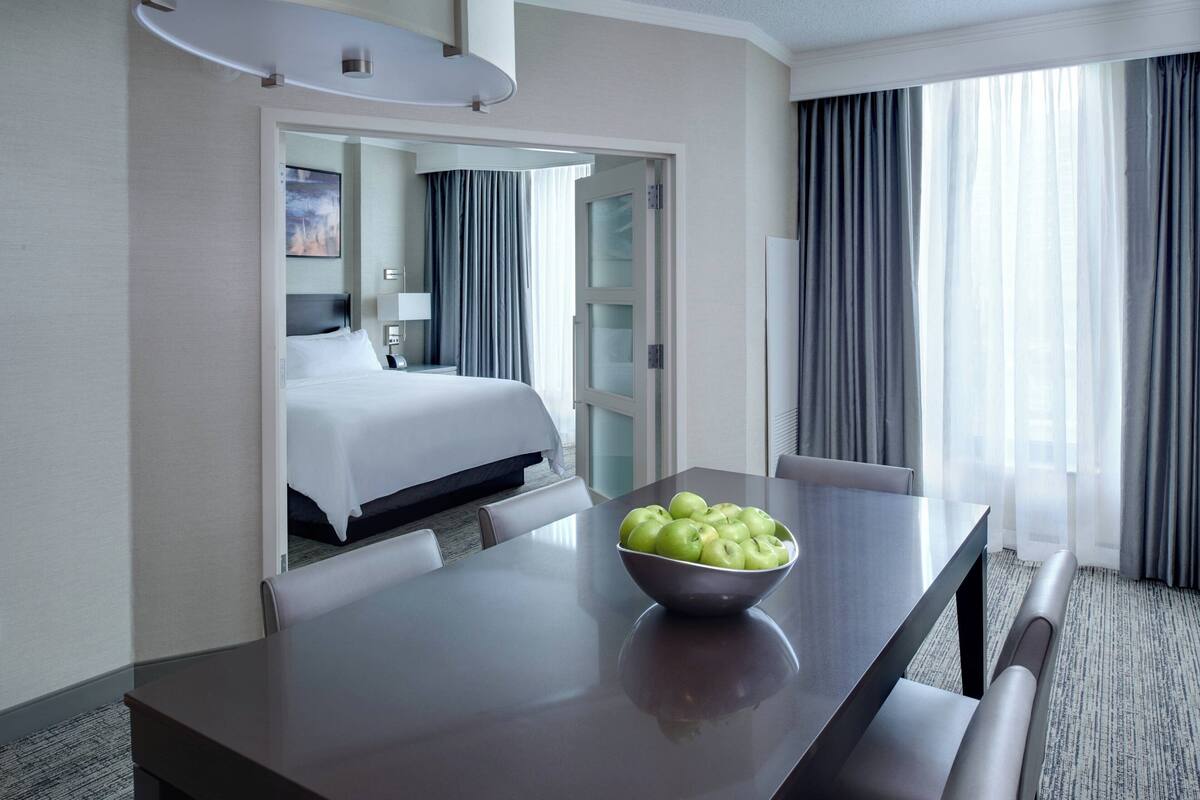A hotel room with a bed, a dining table with a bowl of apples, a mirror, and a view of the outside through large windows.
