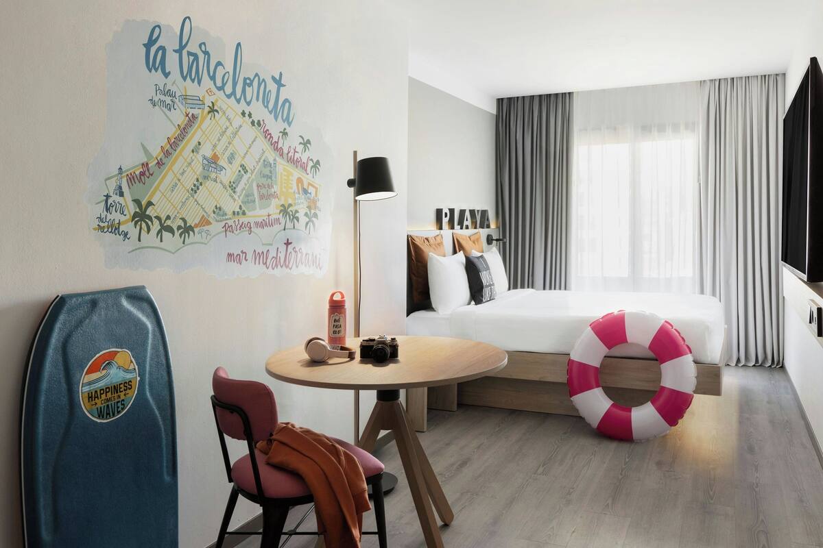 A hotel room with a bed, a round table, a chair, a life preserver, a map on the wall, and a surfboard.