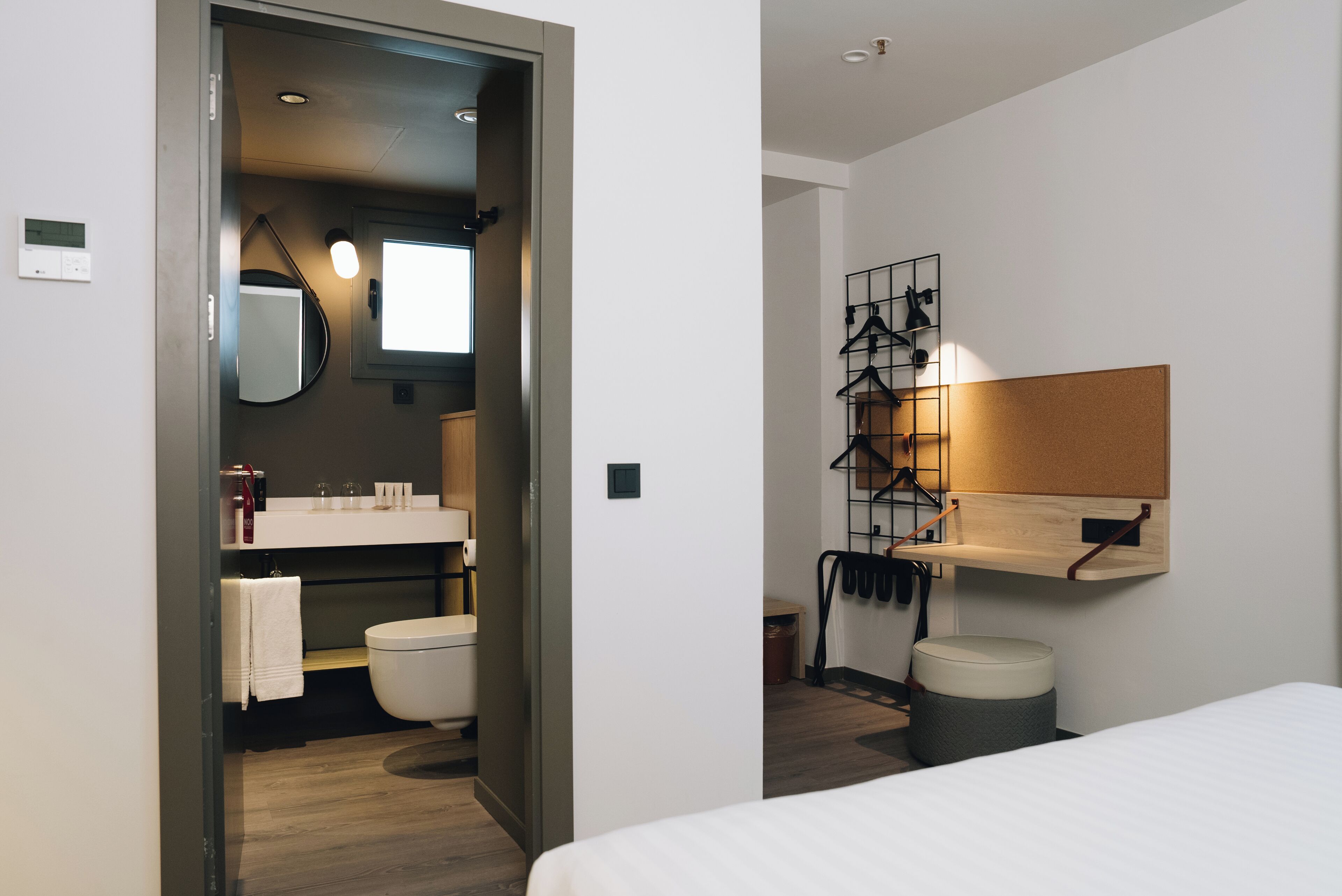 A modern hotel room with a wooden floor, a white bed, a bathroom with a toilet, sink, and mirror, and a wall-mounted shelf with clothes hangers.