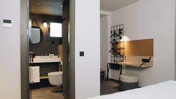 A modern hotel room with a wooden floor, a white bed, a bathroom with a toilet, sink, and mirror, and a wall-mounted shelf with clothes hangers.