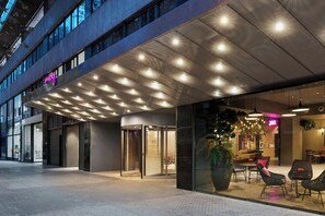 Una entrada de hotel moderna con fachada de vidrio, un letrero de neón y un área de estar al aire libre con muebles de mimbre.