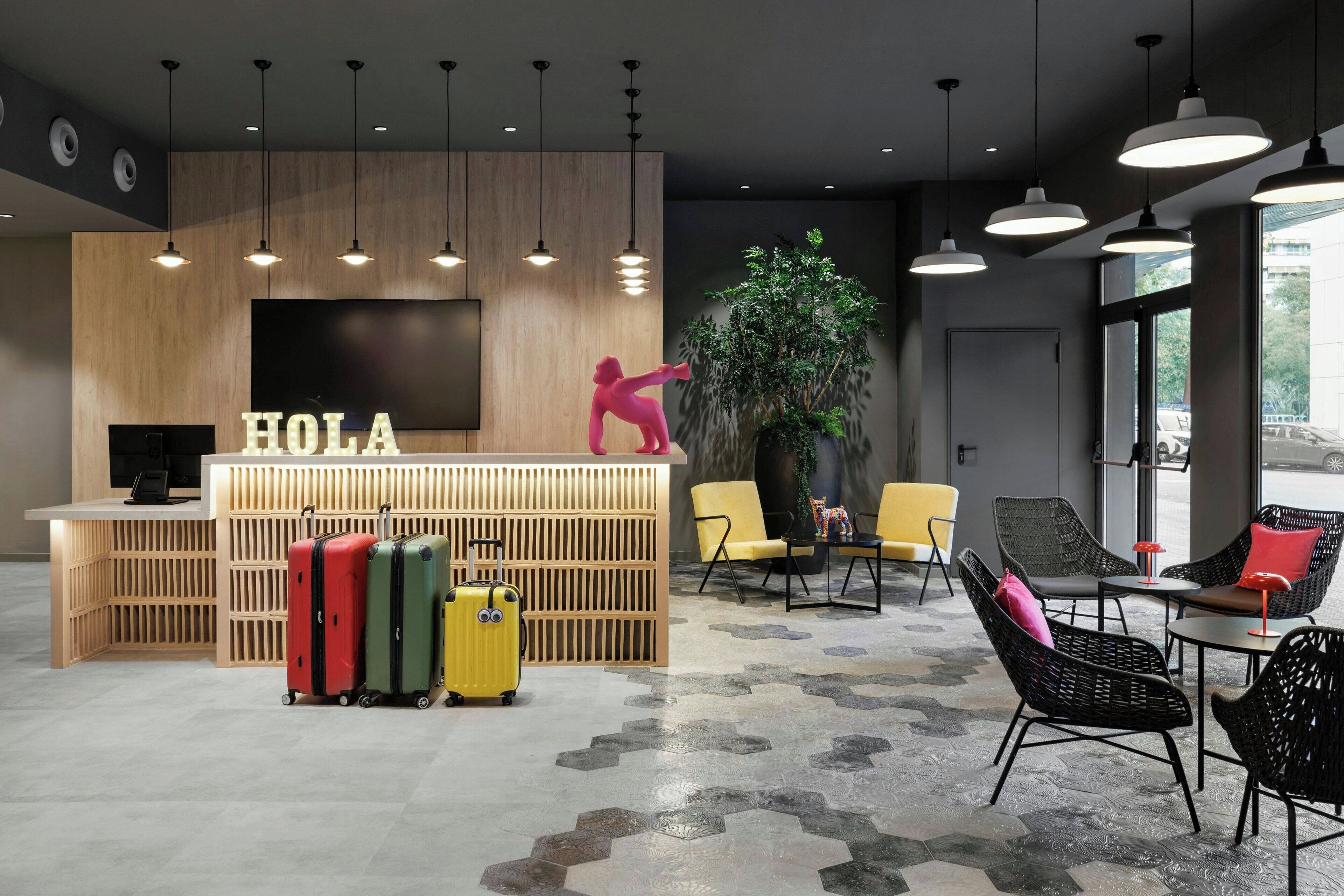 Una zona de recepción moderna con un escritorio de madera, un letrero con la palabra 'HOLA' y tres maletas al frente. Se observan sillas y mesas al fondo, así como una planta grande.