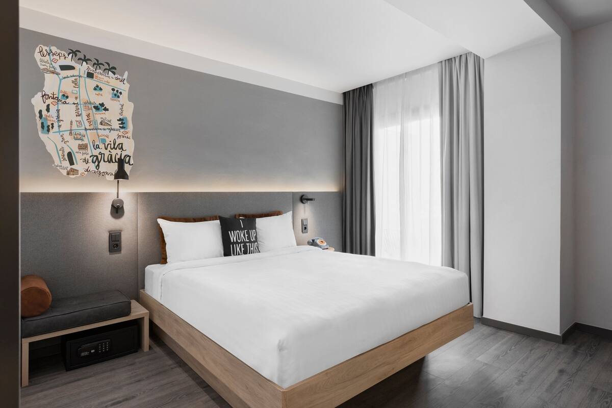 A modern hotel room with a large bed, a bedside table, a wall-mounted artwork, and a window with curtains.
