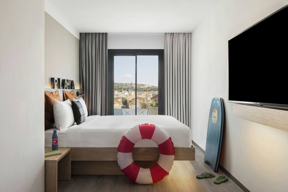 A hotel room with a bed, a view of the city, a TV, and a life preserver on the floor.