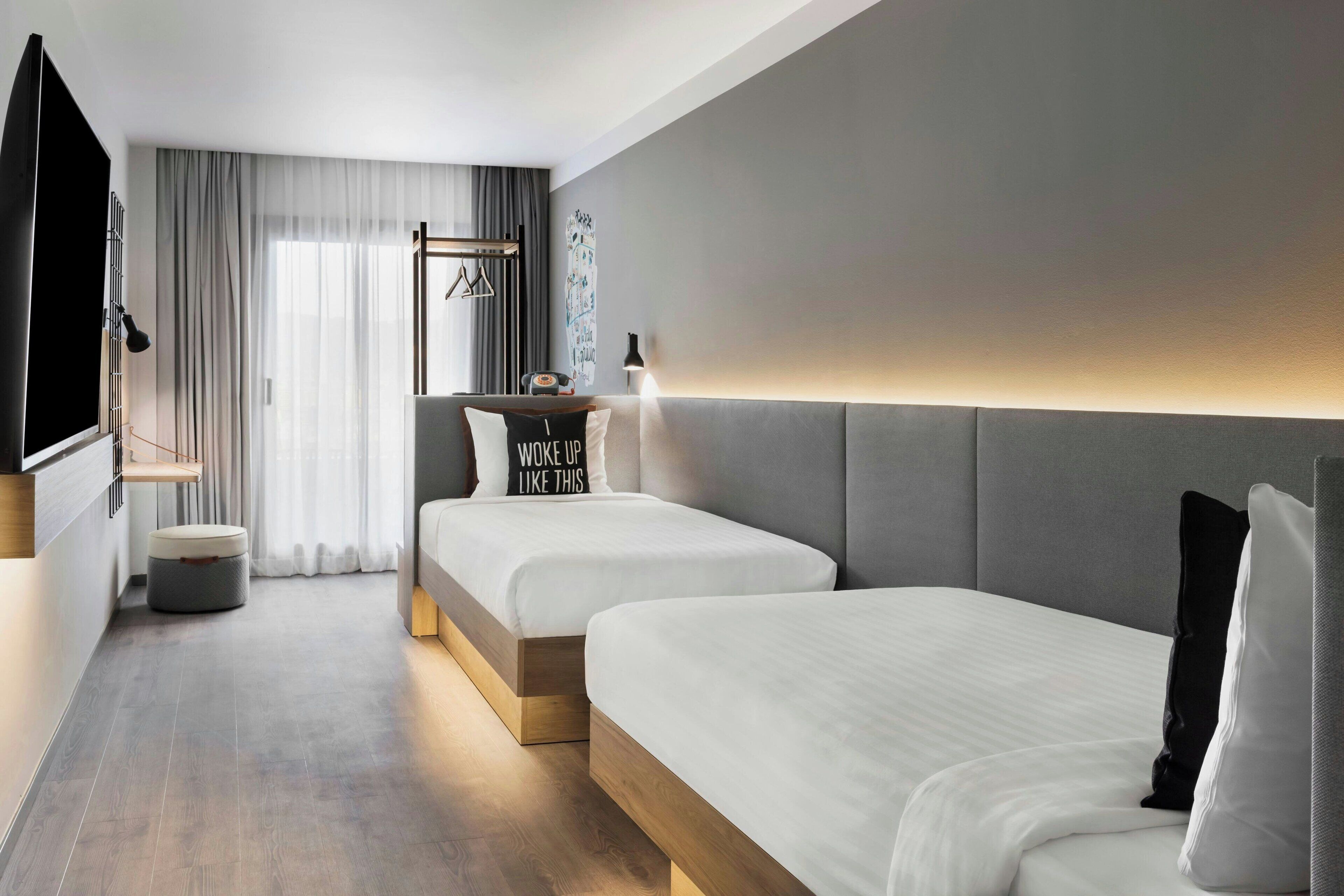 Une chambre d’hôtel moderne avec deux lits, une petite table avec une lampe, un téléviseur et une grande fenêtre avec des rideaux.