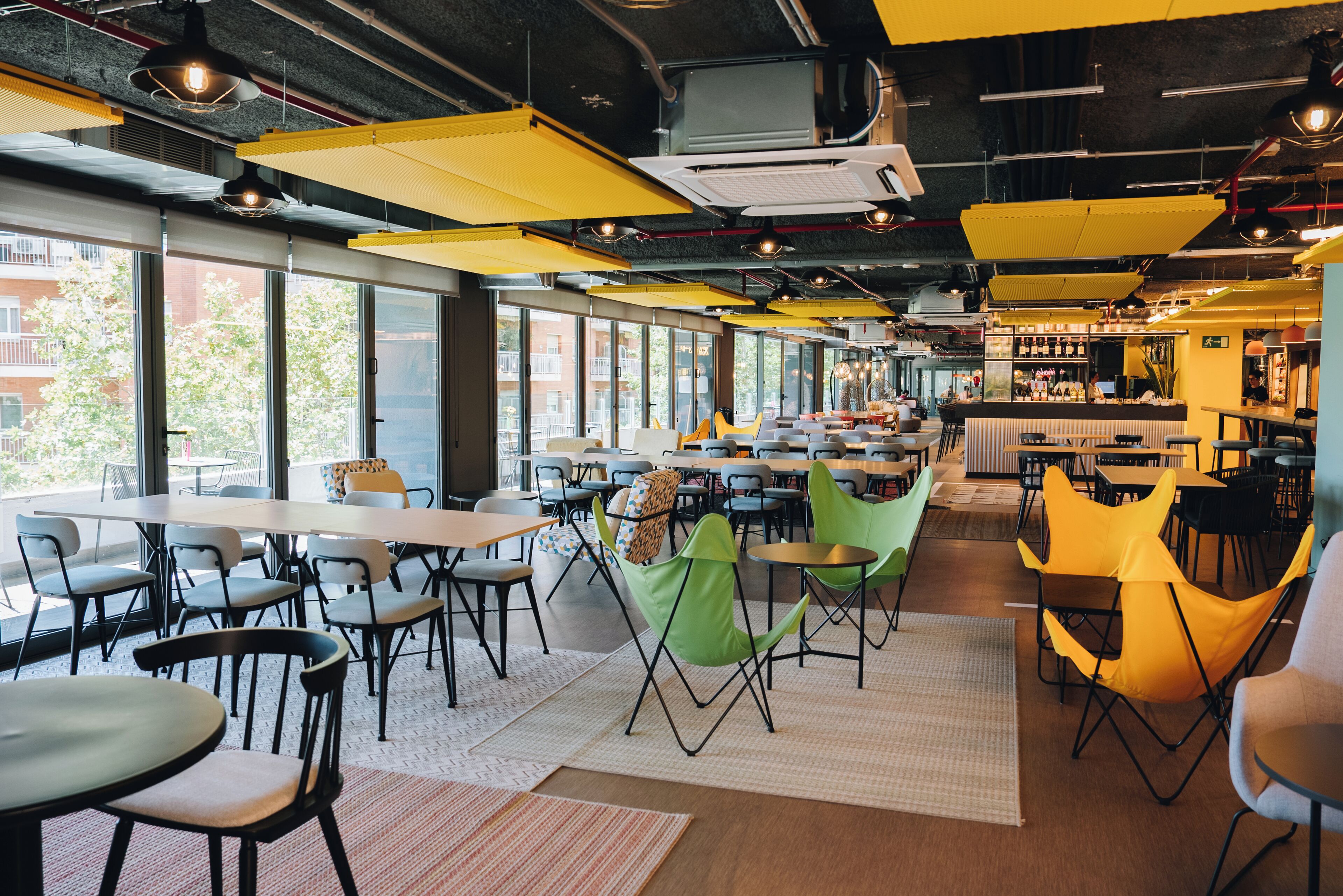 A modern restaurant with high ceilings, yellow hanging lights, and large windows offering views of the city.