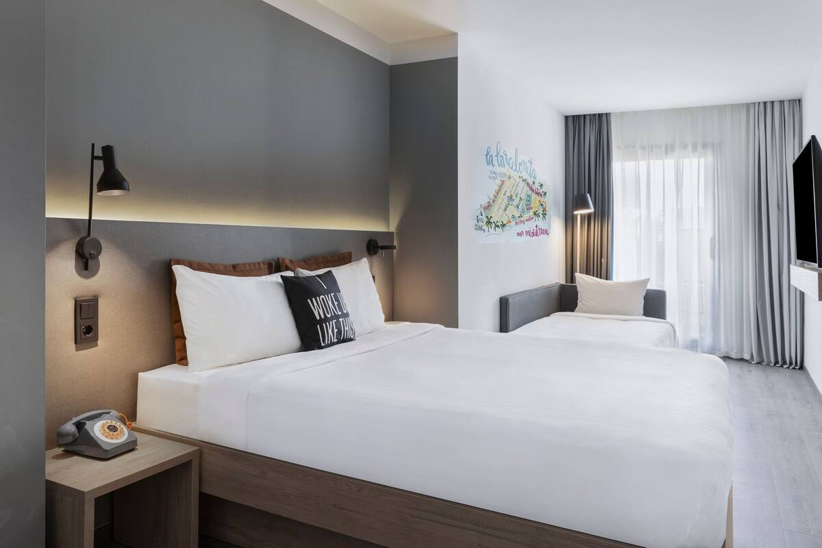 A hotel room with two beds, a bedside table with a telephone, and a wall-mounted light.
