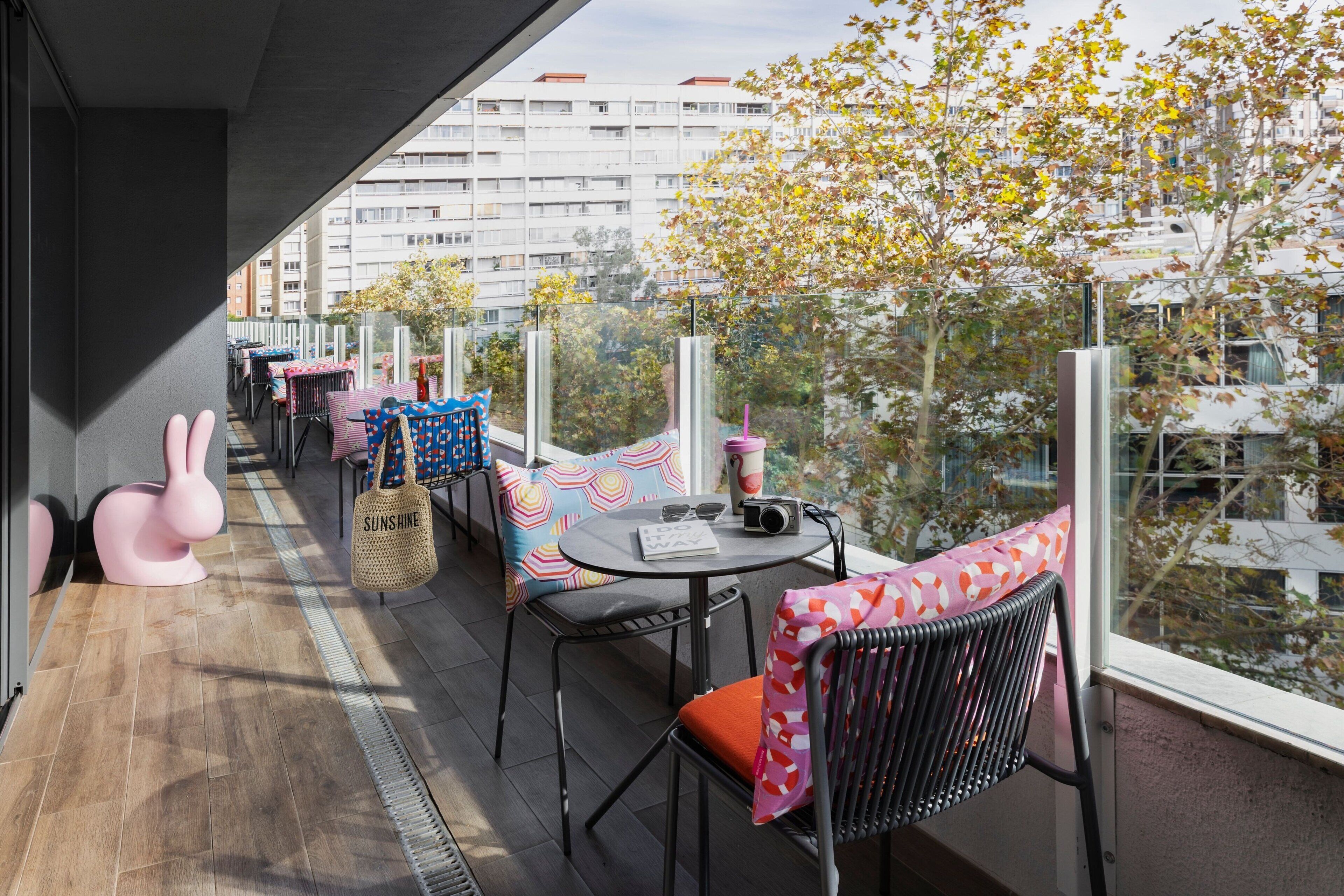 Un balcon avec une rangée de tables et de chaises, une sculpture de lapin rose et une vue sur des bâtiments et des arbres.