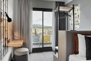 A modern hotel room with a balcony, a hanging lamp, a small wooden shelf, and a bed with pillows. - Moxy Barcelona (Barcelona)
