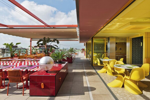 Un espace extérieur aménagé pour les repas, avec des tables et des chaises rouges et jaunes, un barbecue et des plantes en pot.