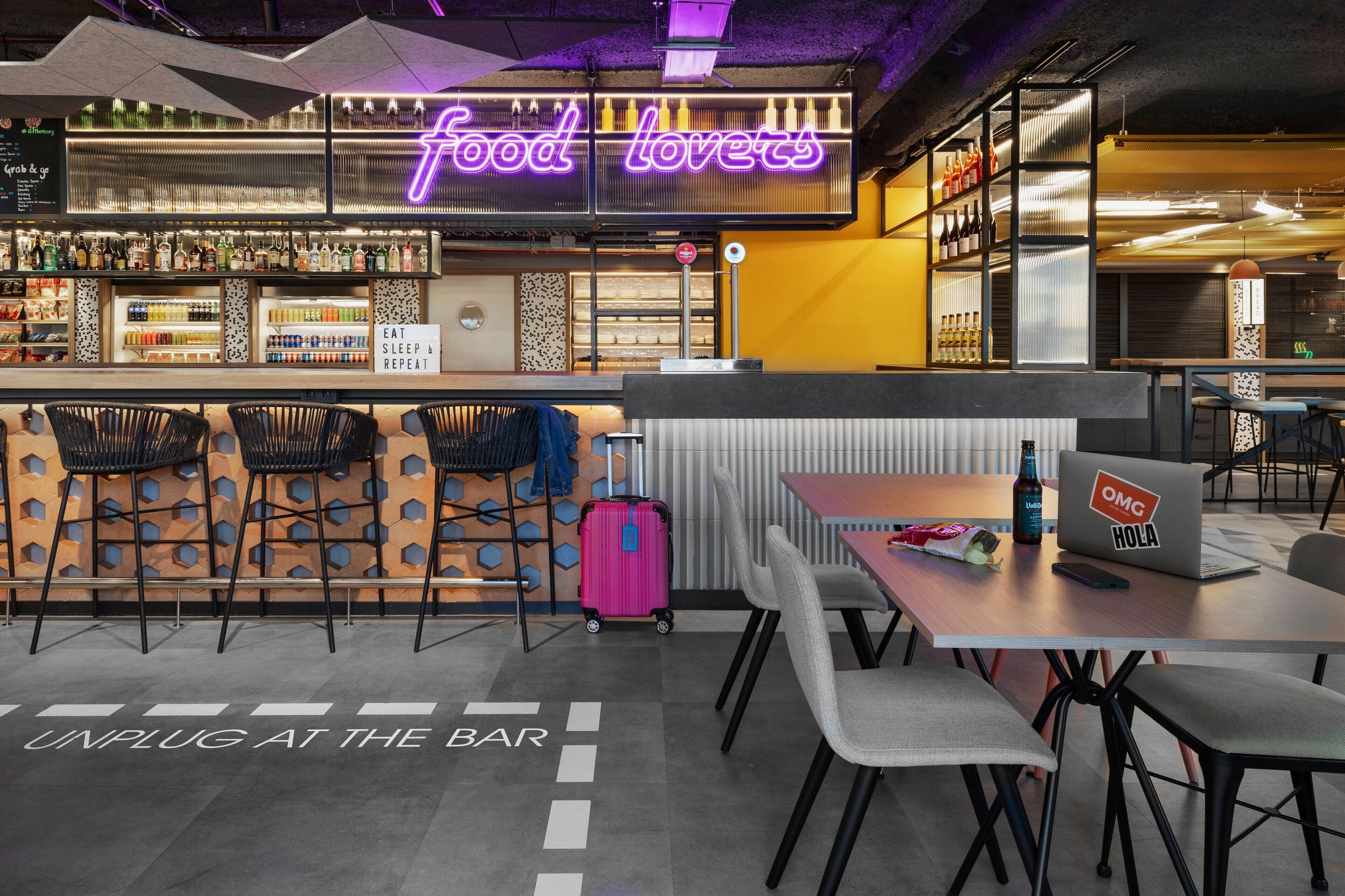 Eine moderne Bar mit einer Neonreklame mit dem Schriftzug "food lovers", Barhocker, einem Laptop und einem Koffer auf einem Tisch.
