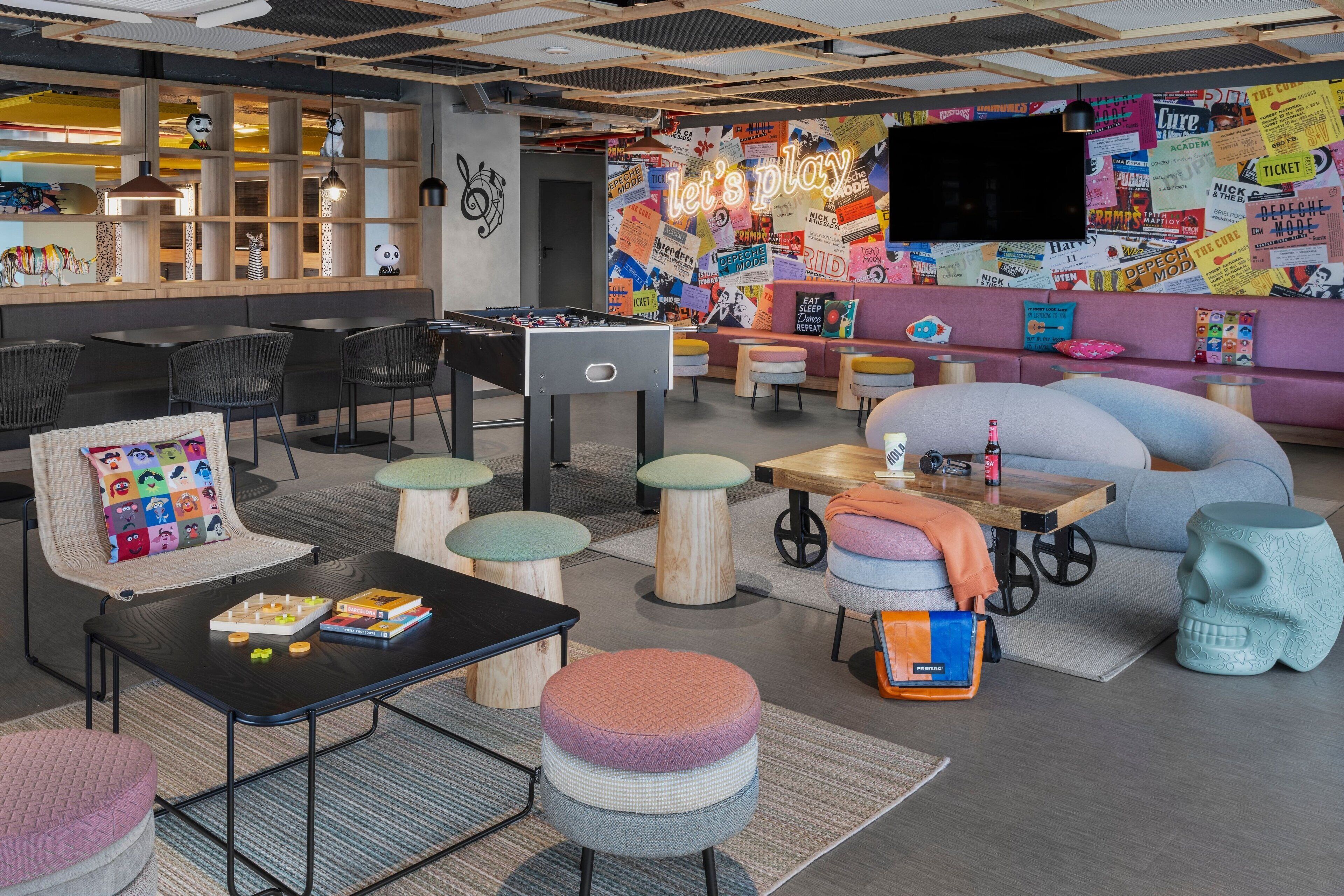 A modern lounge area with seating arrangements, a foosball table, and a wall decorated with various posters and signs.