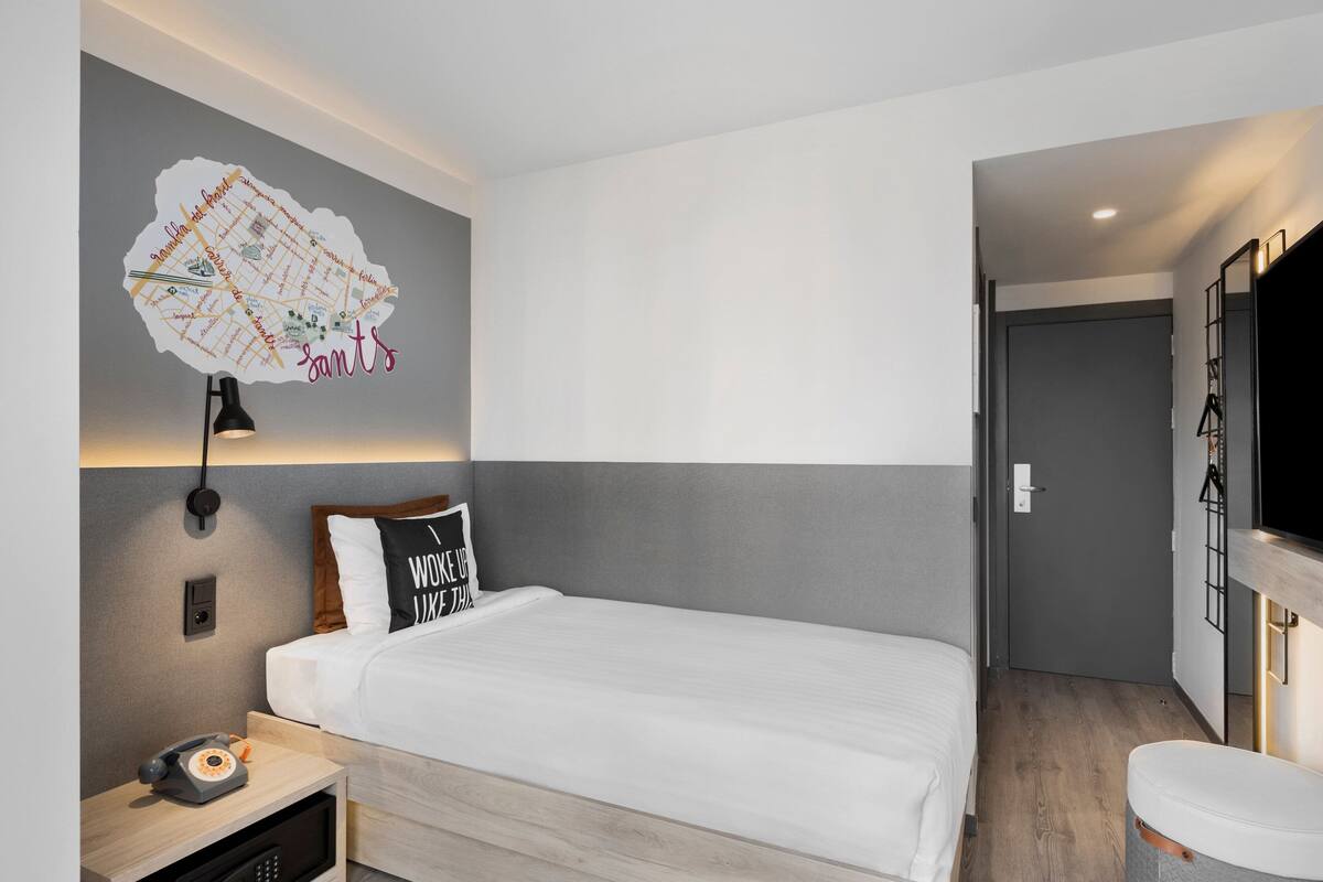 A modern hotel room with a neatly made bed, a wall-mounted map, a bedside lamp, a telephone, and a small desk with a lamp.