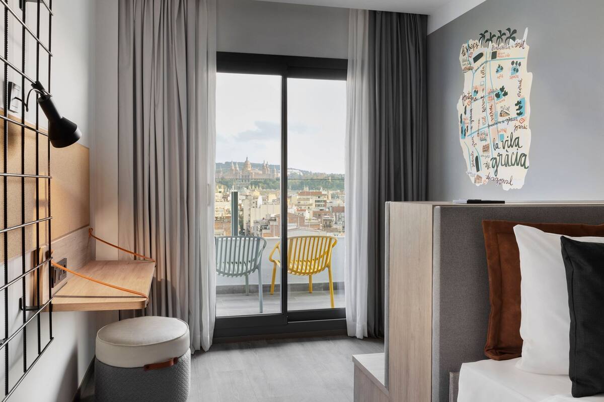 A modern hotel room with a balcony, a city view, a bed with pillows, and a wall art piece.