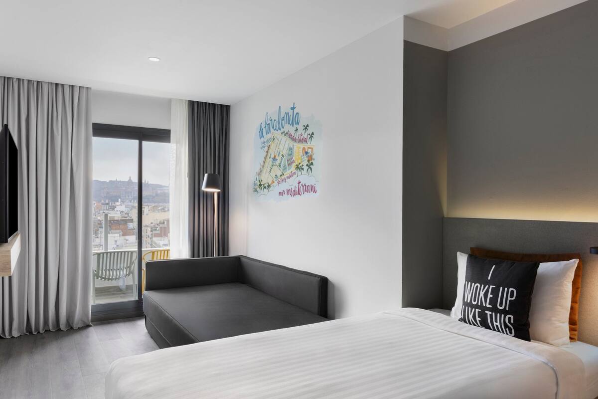 A modern hotel room with a large bed, a sofa, a city view, and a wall mural.